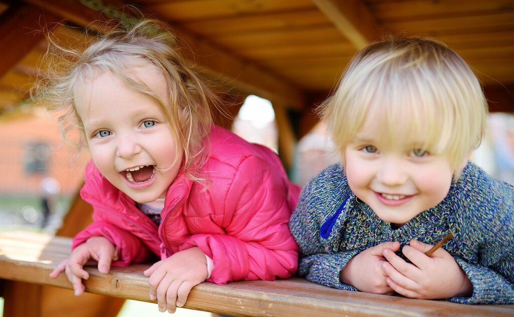 Foto: Zwei kleine Kinder schauen lachend aus einem Holzgerüst auf einem Spielplatz