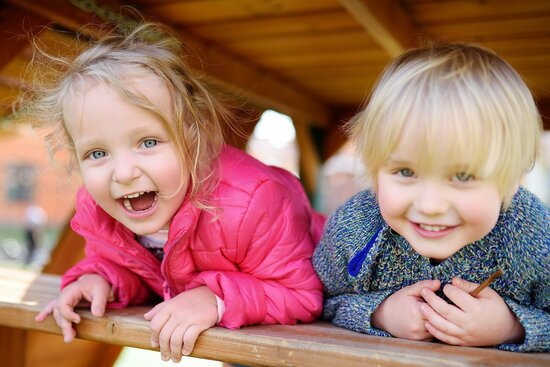 Foto: Zwei kleine Kinder schauen lachend aus einem Holzgerüst auf einem Spielplatz